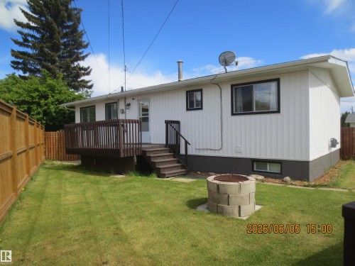Rear exterior featuring white vertical siding, a raised wood deck with railing and stairs, a privacy fence, a grass lawn, and a concrete block fire pit - 1102 55 Street, Edson, AB 