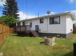Rear exterior featuring white vertical siding, a raised wood deck with railing and stairs, a privacy fence, a grass lawn, and a concrete block fire pit - 
