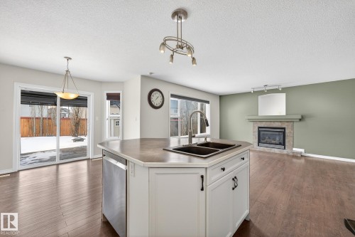 Kitchen featuring a kitchen island with sink, open floor plan, a tiled fireplace, dishwasher, and rail lighting - 152 Rue Marquet, Beaumont, AB - Indoor Photo Showing Kitchen With Fireplace With Double Sink