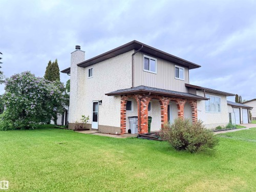 Exterior showcasing a combination of stucco and vertical siding, a covered arched brick entryway, multiple windows, an attached garage, and a lush green lawn - 10503 110 Street, Westlock, AB 