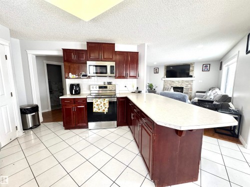 Kitchen featuring stainless steel appliances, a stone fireplace, light countertops, a peninsula, and reddish brown cabinets - 6734 162A Avenue, Edmonton, AB - Indoor Photo Showing Kitchen With Fireplace