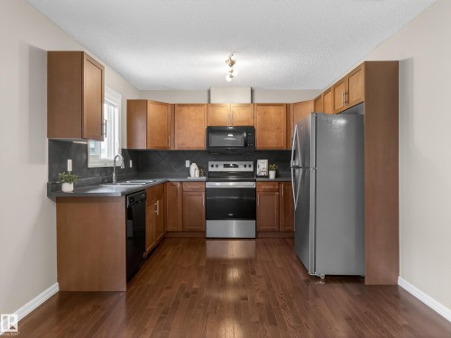 Kitchen with dark countertops, wood finish cabinetry, black appliances, dark wood finished floors, and a textured ceiling - 125 655 Tamarack Road, Edmonton, AB - Indoor Photo Showing Kitchen