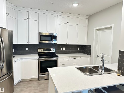 Modern kitchen featuring white shaker-style cabinetry, stainless steel appliances, dark subway tile backsplash, light-toned countertops, and wood-finish flooring - 437 1818 Rutherford Road, Edmonton, AB - Indoor Photo Showing Kitchen With Stainless Steel Kitchen With Double Sink With Upgraded Kitchen