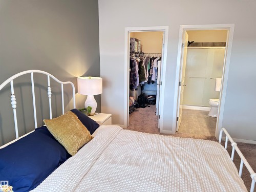 Bedroom featuring a white metal bed frame, nightstand with lamp, carpet flooring, and direct access to a full bathroom with a glass-enclosed shower and a separate closet with built-in shelving - 437 1818 Rutherford Road, Edmonton, AB - Indoor Photo Showing Bedroom