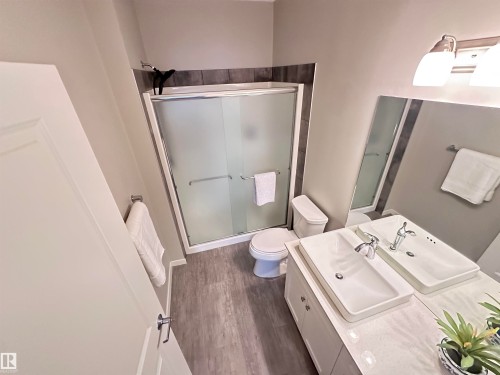 Bathroom featuring a double vanity with rectangular vessel sinks, a framed mirror, a two-light vanity fixture, a shower with a sliding frosted glass door, and wood-finish flooring - 437 1818 Rutherford Road, Edmonton, AB - Indoor Photo Showing Bathroom