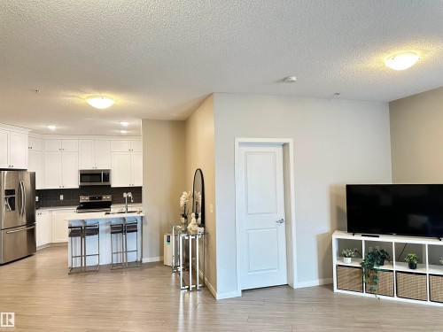 Open-concept living area featuring light wood-finish flooring, white flat-panel cabinetry, stainless steel appliances, a dark subway tile backsplash, and a kitchen island with a sink - 437 1818 Rutherford Road, Edmonton, AB - Indoor Photo Showing Kitchen