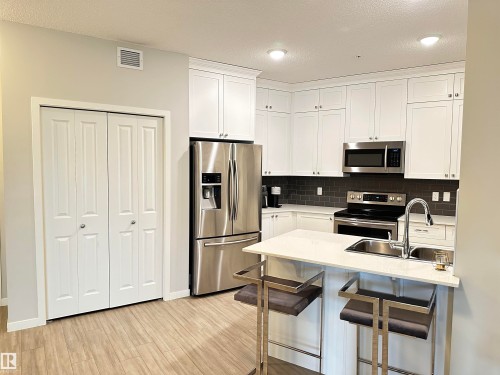 Modern kitchen featuring white shaker cabinetry, stainless steel appliances, a dark subway tile backsplash, light stone countertops, and wood-finish flooring - 437 1818 Rutherford Road, Edmonton, AB - Indoor Photo Showing Kitchen With Stainless Steel Kitchen With Double Sink