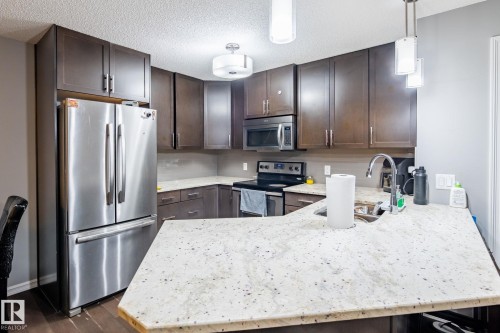 Kitchen with dark wood finish cabinetry, stainless steel appliances, light stone countertops, a peninsula, and a textured ceiling - 207 2203 44 Avenue, Edmonton, AB - Indoor Photo Showing Kitchen With Stainless Steel Kitchen