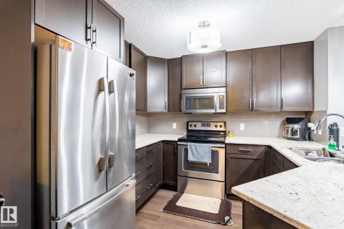 Kitchen featuring stainless steel appliances, dark wood finish cabinets, a textured ceiling, light stone counters, and light wood-type flooring - 207 2203 44 Avenue, Edmonton, AB - Indoor Photo Showing Kitchen With Stainless Steel Kitchen With Upgraded Kitchen