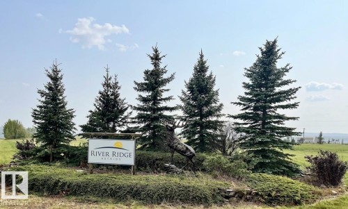 Community entrance sign for River Ridge, featuring five mature evergreen trees, a stone-lined planter with low-lying shrubbery, and open green space - 30 River Ridge Estates, Rural Wetaskiwin County, AB 