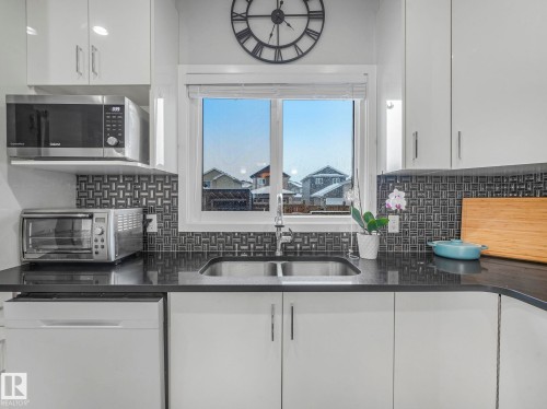 Kitchen featuring white cabinets, white dishwasher, stainless steel microwave, dark stone counters, and backsplash - 3510 48 Avenue, Beaumont, AB - Indoor Photo Showing Kitchen With Double Sink