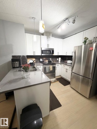 Kitchen with stainless steel appliances, white cabinets, hanging light fixtures, a breakfast bar, and a peninsula - 706 5151 Windermere Bv, Edmonton, AB - Indoor Photo Showing Kitchen With Double Sink
