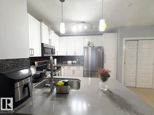 Kitchen with white cabinets, stainless steel appliances, decorative light fixtures, a textured ceiling, and dark stone countertops - 706 5151 Windermere Bv, Edmonton, AB - Indoor Photo Showing Kitchen With Double Sink With Upgraded Kitchen