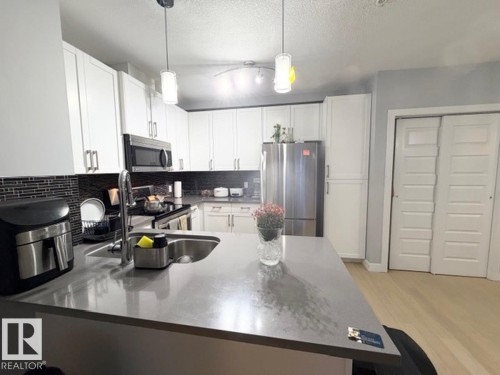 Kitchen featuring white cabinets, a peninsula, stainless steel appliances, light wood finished floors, and hanging light fixtures - 706 5151 Windermere Bv, Edmonton, AB - Indoor Photo Showing Kitchen With Double Sink With Upgraded Kitchen