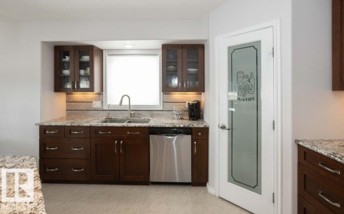 Kitchen with dark wood finish cabinetry, dishwasher, light stone countertops, decorative backsplash, and glass fronted cabinets - 11116 103 Street, Westlock, AB - Indoor Photo Showing Kitchen With Double Sink