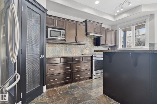 Kitchen with stainless steel appliances, a breakfast bar area, dark wood finish cabinets, decorative backsplash, and dark stone counters - 613 Stewart Crescent, Edmonton, AB - Indoor Photo Showing Kitchen