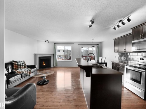 Kitchen featuring open floor plan, stainless steel appliances, a center island with sink, rail lighting, and a warm lit fireplace - 1716 64 Street, Edmonton, AB - Indoor Photo Showing Kitchen With Fireplace
