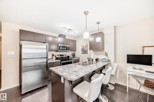 Kitchen with stainless steel appliances, a breakfast bar area, a peninsula, light countertops, and dark wood-style flooring - 403 340 Windermere Road, Edmonton, AB - Indoor Photo Showing Kitchen With Stainless Steel Kitchen With Double Sink