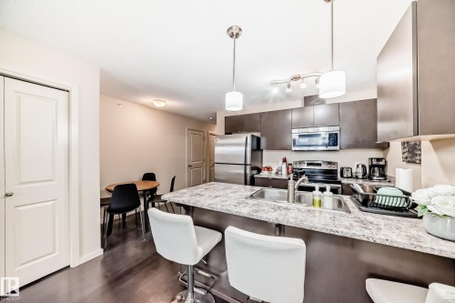Kitchen with a kitchen bar, a peninsula, dark wood finish cabinetry, stainless steel appliances, and hanging light fixtures - 403 340 Windermere Road, Edmonton, AB - Indoor Photo Showing Kitchen With Stainless Steel Kitchen With Upgraded Kitchen