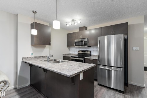 Kitchen featuring stainless steel appliances, a kitchen breakfast bar, a peninsula, hanging light fixtures, and a textured ceiling - 231 344 Windermere Road, Edmonton, AB - Indoor Photo Showing Kitchen With Double Sink