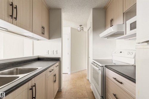 Kitchen with dark countertops, white appliances, a textured ceiling, and light wood finish cabinetry - 312 11620 9A Avenue, Edmonton, AB - Indoor Photo Showing Kitchen With Double Sink