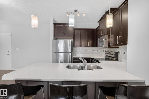 Kitchen with a peninsula, stainless steel appliances, a breakfast bar area, dark wood finish cabinets, and backsplash - 206 812 Welsh Drive, Edmonton, AB - Indoor Photo Showing Kitchen With Stainless Steel Kitchen With Double Sink With Upgraded Kitchen