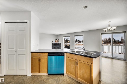 Kitchen with dark countertops, dishwasher, a peninsula, suspended lighting, and a tiled fireplace - 3401 47 Street, Beaumont, AB - Indoor Photo Showing Kitchen With Double Sink