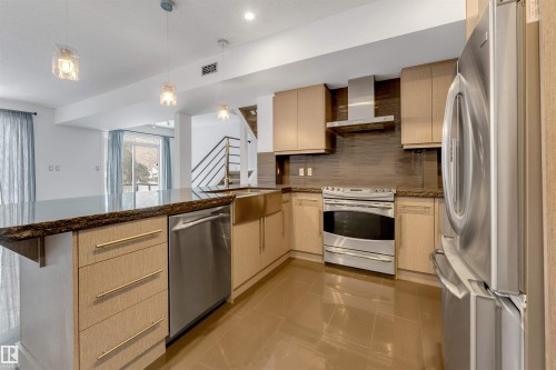 Kitchen featuring dark stone countertops, light wood finish cabinets, stainless steel appliances, a peninsula, and decorative light fixtures - 102 10711 83 Avenue, Edmonton, AB - Indoor Photo Showing Kitchen With Stainless Steel Kitchen