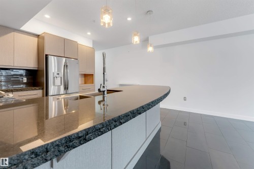 Kitchen featuring stainless steel refrigerator with ice dispenser, decorative light fixtures, light wood finish cabinetry, dark stone countertops, and decorative backsplash - 102 10711 83 Avenue, Edmonton, AB - Indoor Photo Showing Kitchen