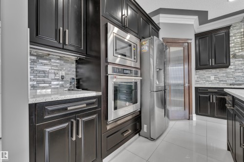 Kitchen featuring decorative backsplash, stainless steel appliances, light stone countertops, crown molding, and light tile patterned floors - 2612 Adam Court Sw, Edmonton, AB - Indoor Photo Showing Kitchen With Upgraded Kitchen