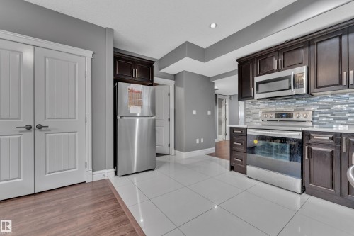 Kitchen with dark wood finish cabinetry, stainless steel appliances, light tile patterned floors, tasteful backsplash, and light stone countertops - 2612 Adam Court Sw, Edmonton, AB - Indoor Photo Showing Kitchen