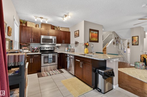 Kitchen featuring a peninsula, stainless steel appliances, light tile patterned flooring, backsplash, and dark wood finish cabinetry - 14 1140 Chappelle Boulevard, Edmonton, AB - Indoor Photo Showing Kitchen With Double Sink