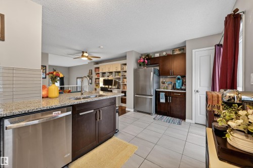 Kitchen featuring stainless steel appliances, dark wood finish cabinetry, light tile patterned floors, ceiling fan, and light stone countertops - 14 1140 Chappelle Boulevard, Edmonton, AB - Indoor Photo Showing Kitchen With Double Sink