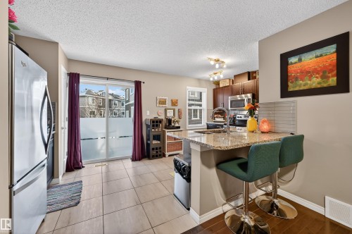 Kitchen featuring a kitchen bar, stainless steel appliances, a peninsula, light stone counters, and a textured ceiling - 14 1140 Chappelle Boulevard, Edmonton, AB - Indoor