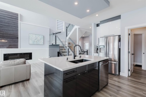 Kitchen featuring modern cabinets, open floor plan, stainless steel appliances, an island with sink, and a glass covered fireplace - 16217 32 Avenue, Edmonton, AB - Indoor Photo Showing Kitchen With Double Sink With Upgraded Kitchen
