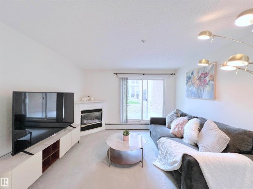 Living room with a glass covered fireplace, a baseboard heating unit, light carpet, and a textured ceiling - 110 10511 42 Avenue, Edmonton, AB - Indoor Photo Showing Living Room With Fireplace