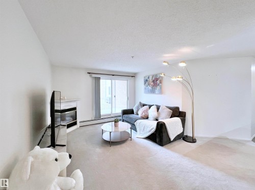 Living room featuring light carpet, a textured ceiling, baseboard heating, and a high end fireplace - 110 10511 42 Avenue, Edmonton, AB - Indoor Photo Showing Living Room With Fireplace