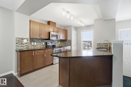 Kitchen with stainless steel appliances, a peninsula, dark stone counters, a textured ceiling, and decorative backsplash - 71 4029 Orchards Drive, Edmonton, AB - Indoor Photo Showing Kitchen