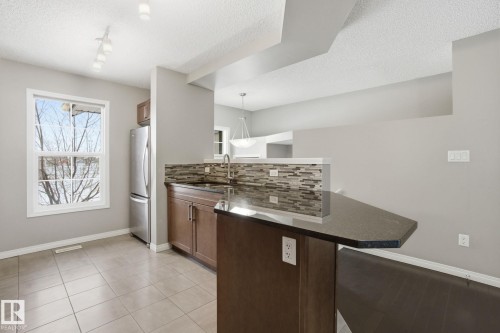 Kitchen featuring a peninsula, backsplash, freestanding refrigerator, dark stone counters, and a textured ceiling - 71 4029 Orchards Drive, Edmonton, AB - Indoor Photo Showing Kitchen