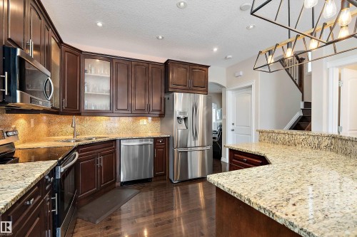 Kitchen featuring stainless steel appliances, dark wood finish cabinetry, glass fronted cabinets, light stone countertops, and decorative light fixtures - 10623 75 Avenue, Edmonton, AB - Indoor Photo Showing Kitchen With Upgraded Kitchen