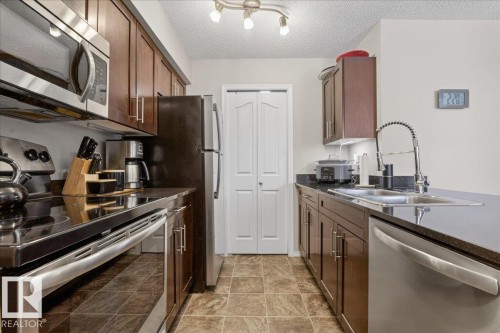 Kitchen with stainless steel appliances, a textured ceiling, dark wood finish cabinetry, and dark stone counters - 305 12045 22 Avenue, Edmonton, AB - Indoor Photo Showing Kitchen With Double Sink