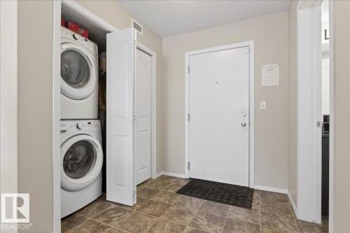 Laundry area featuring a textured ceiling, stacked washer / drying machine, and stone finish flooring - 305 12045 22 Avenue, Edmonton, AB - Indoor Photo Showing Laundry Room