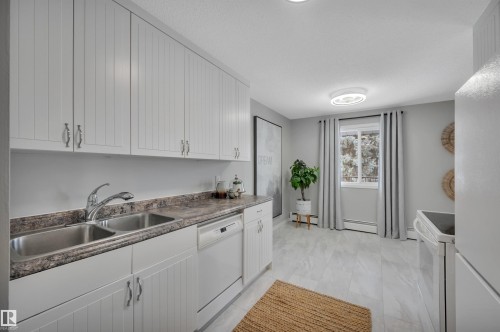 Kitchen with white appliances, white cabinets, dark countertops, and baseboard heating - 201 237 Woodvale Road W, Edmonton, AB - Indoor Photo Showing Kitchen With Double Sink