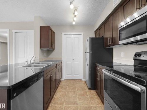 Kitchen with stainless steel appliances, a peninsula, a textured ceiling, dark wood finish cabinets, and track lighting - 422 5816 Mullen Place, Edmonton, AB - Indoor Photo Showing Kitchen With Double Sink