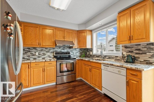 Kitchen featuring appliances with stainless steel finishes, dark wood-type flooring, light stone counters, brown cabinets, and under cabinet range hood - 11608 11A Avenue, Edmonton, AB - Indoor Photo Showing Kitchen With Double Sink