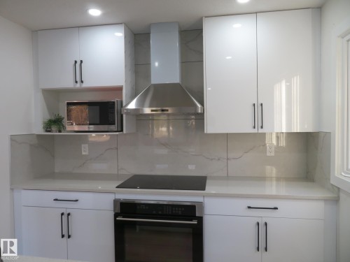 Modern kitchen featuring glossy white cabinetry with matte black hardware, stone countertops, a tiled backsplash with subtle veining, a stainless steel range hood, and a built-in oven - 13520 113A Street, Edmonton, AB - Indoor Photo Showing Kitchen