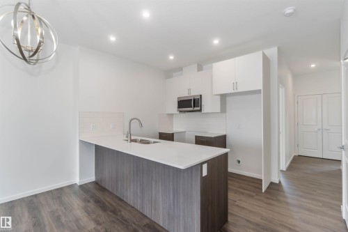 Dual tone kitchen featuring a peninsula, tasteful backsplash, dark wood-style flooring, stainless steel microwave, and light stone countertops - 2467 Alces Link Link, Edmonton, AB - Indoor Photo Showing Kitchen With Double Sink