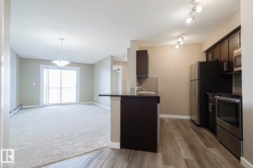 This open-concept living space features light-colored carpeting, a sliding glass door providing natural light, and a modern pendant light fixture - 313 1510 Watt Drive, Edmonton, AB - Indoor Photo Showing Kitchen With Stainless Steel Kitchen