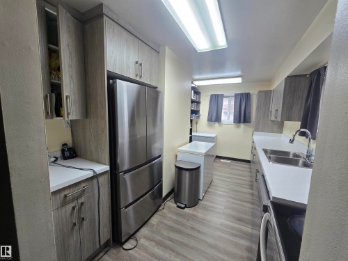 Galley kitchen featuring wood-finish cabinetry, stainless steel refrigerator, white countertops, double basin sink, and wood-finish flooring - 2 4403 Riverbend Road, Edmonton, AB 