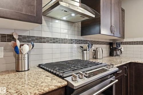Kitchen featuring dark wood finish cabinetry, exhaust hood, stainless steel gas stove, light stone counters, and decorative backsplash - 2031 Westerra Loop, Stony Plain, AB - Indoor Photo Showing Kitchen With Upgraded Kitchen